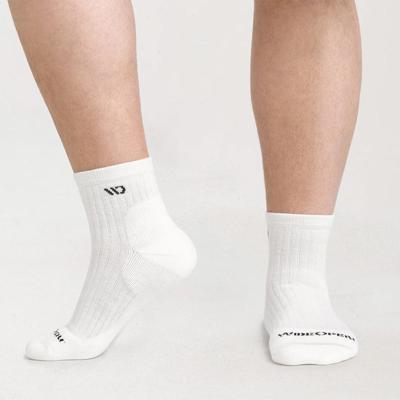 White crew socks worn on a person's feet, shown against a clean light gray studio background. The socks have black printed branding near the toe and ankle.