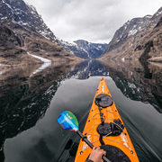 Orange kayak with a multicolor paddle floating down a calm river with snowy mountains on either side and in the background.