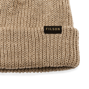 Close up of a sawdust brown beanie with a tan Filson logo on it against a white background. 