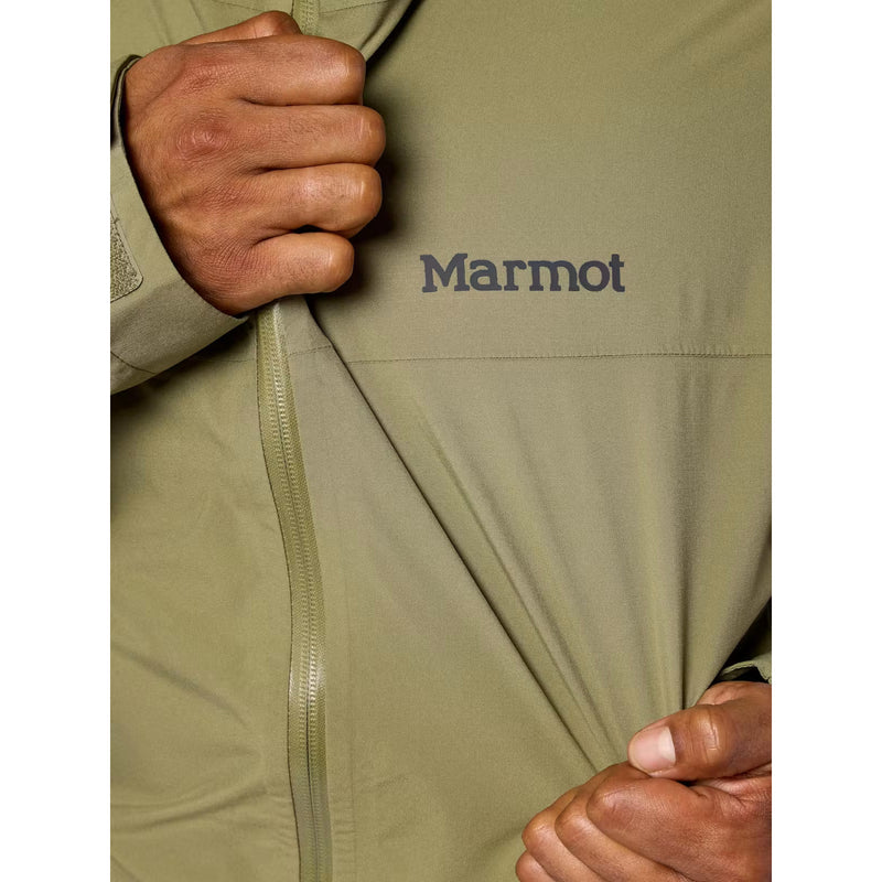 Close-up of a person wearing a Marmot jacket in olive green, pulling the fabric slightly to show the zipper and the Marmot logo on the chest.