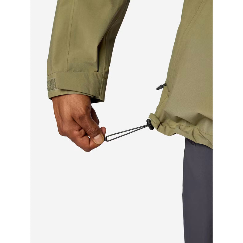 Close-up of a person's hand adjusting the drawstring closure on the hem of an olive green jacket, with dark gray pants visible underneath, set against a plain light gray background.