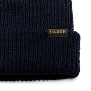 Close up of a navy blue beanie with a tan Filson logo against a white background. 
