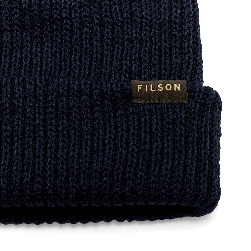 Close up of a navy blue beanie with a tan Filson logo against a white background. 