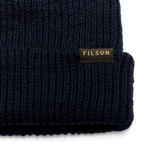 Close up of a navy blue beanie with a tan Filson logo against a white background. 