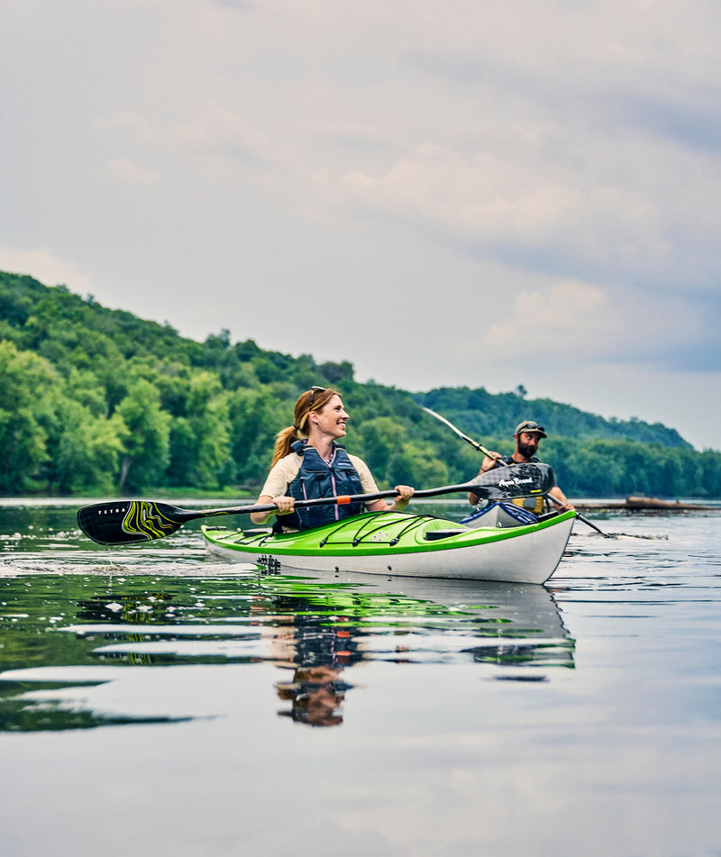 Woman paddling her green and white kayak in a calm lake next to a man in a blue and white kayak with trees in the background.
