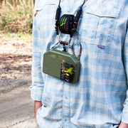 A close-up of a person wearing a light blue plaid shirt outdoors, carrying a green tackle box on a strap with fishing rods and a fly fishing lure attached.