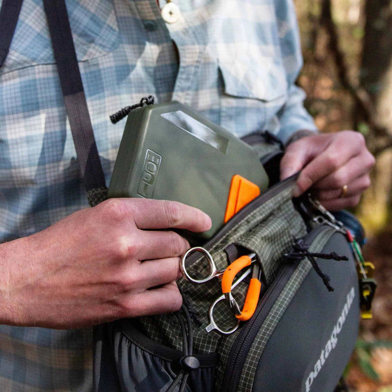 Person holding a green container and black bag with outdoor equipment in a forest setting