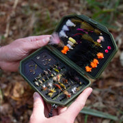 Hand holding a green fly fishing lure box with various colorful flies against a natural background.