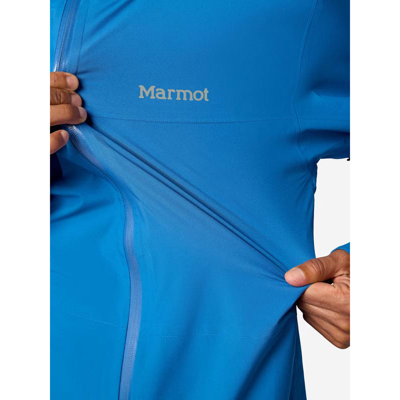 Close-up of a person wearing a bright blue Marmot jacket, pulling the fabric to show its stretch, with the Marmot logo visible on the chest.