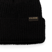 Close up of a black beanie with a tan Filson logo on it against a white background.
