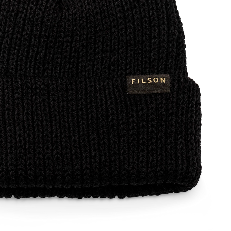 Close up of a black beanie with a tan Filson logo on it against a white background.
