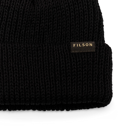 Close up of a black beanie with a tan Filson logo on it against a white background.