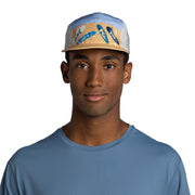 A man wearing a light blue and beige 5-panel cap with blue surfboard graphics on the front panels, paired with a matching dusty blue t-shirt, against a plain white background.