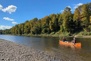 Two men paddling an orange canoe down a river next to a rocky shoreline on a sunny day.