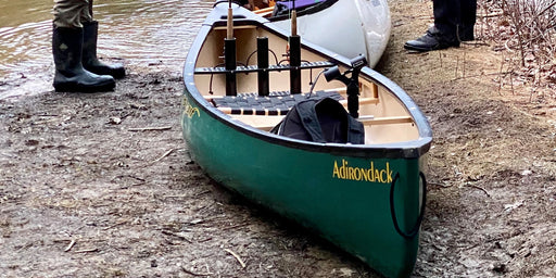 Dark green canoe sitting in the dirt next to a river with people standing in the background.