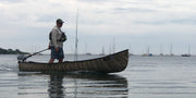 Man standing in a camouflage patterned canoe with fishing gear in a lake with boats sailing in the background. 