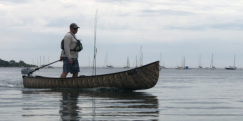 Man standing in a camouflage patterned canoe with fishing gear in a lake with boats sailing in the background. 