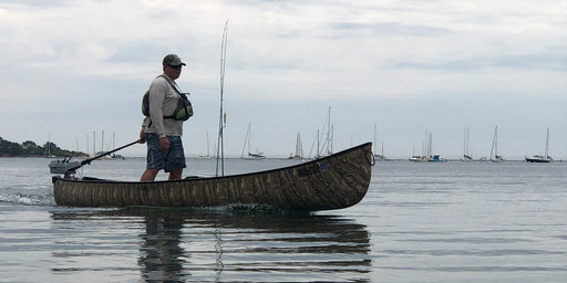 Man standing in a camouflage patterned canoe with fishing gear in a lake with boats sailing in the background. 