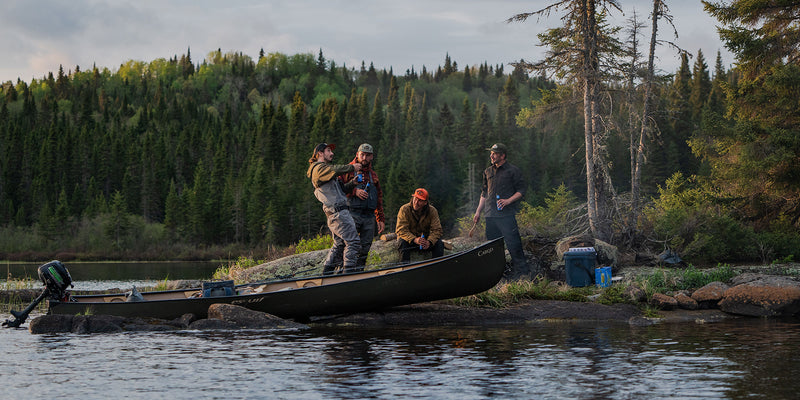 Group of men with fishing poles next to a black canoe in the wilderness.