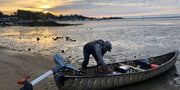 Man checking his fishing gear in a camouflage patterned canoe at the edge of a lake. 