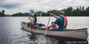 A family paddling their tan canoe in a lake with a scenic view in the background.