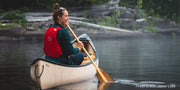 Woman wearing a red life jacket paddling a tan canoe down a calm river.