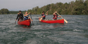 Two red canoes each with two people paddling down a lake with trees in the background.