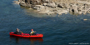 Two women paddling their bright red kayak down a calm looking river with rocks in the background.