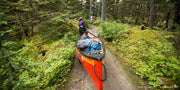 A woman carrying a bright red canoe full of gear and various supplies on a trail through a forest.