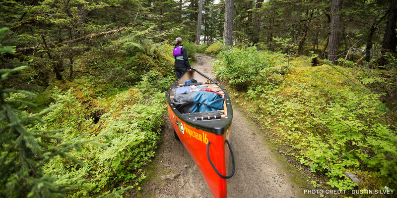 A woman carrying a bright red canoe full of gear and various supplies on a trail through a forest.