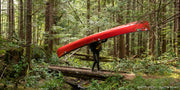 A woman carrying a bright red canoe over a bridge in a thick forest.