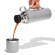 Person pouring coffee from a silver insulated travel mug into a smaller metal cup.
