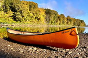 Side view of an orange canoe sitting on a rocky shoreline next to a river on a sunny day with trees in the background.
