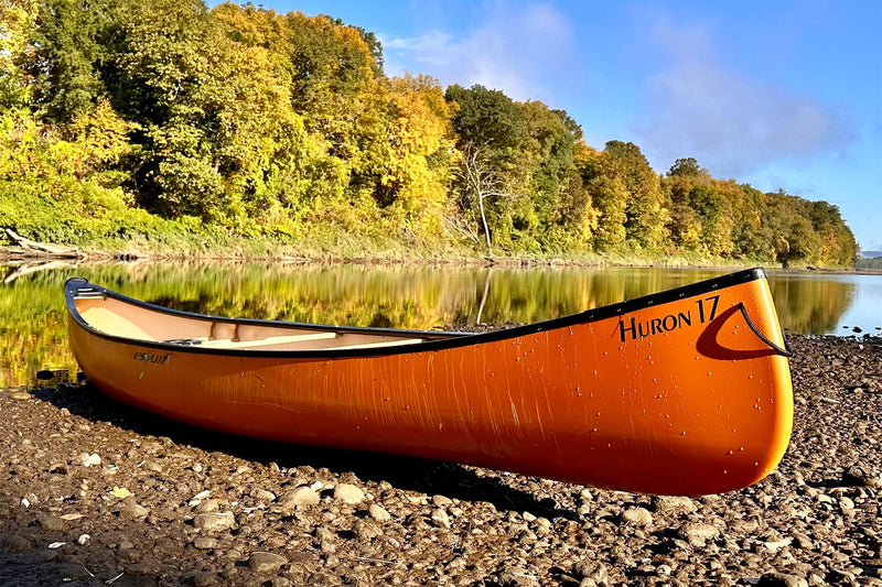 Side view of an orange canoe sitting on a rocky shoreline next to a river on a sunny day with trees in the background.