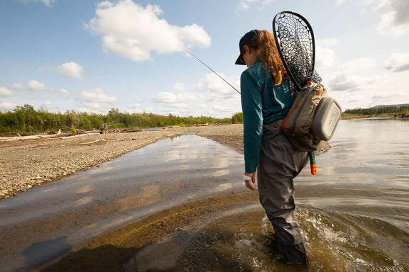 Woman wading through shallow water carrying an assortment of fishing gear with clear skies in the background.