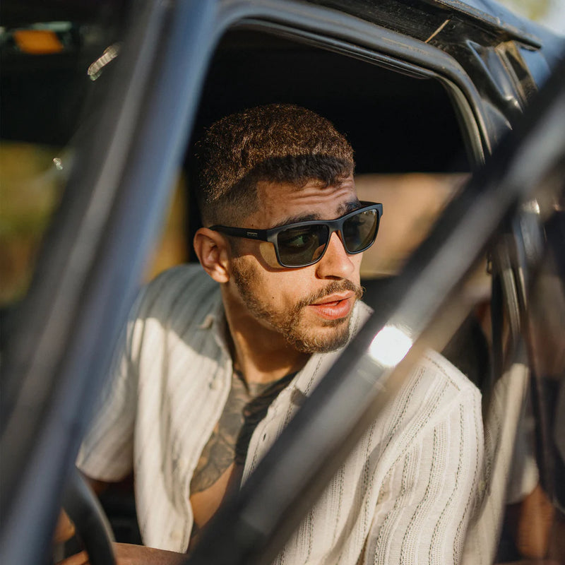 Man sitting in a car wearing a pair of black rimmed sunglasses with dark tinted lenses.