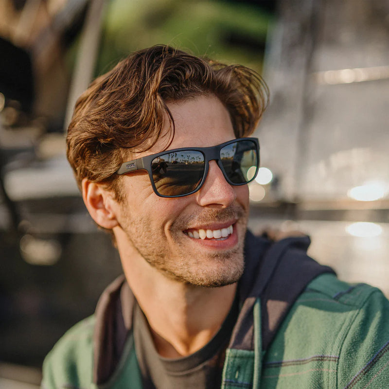 Man wearing a pair of black rimmed sunglasses with dark tinted lenses leaning against a car.