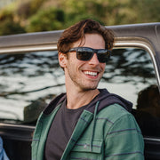 Man standing by a car wearing a pair of black rimmed sunglasses with dark tinted lenses with foliage in the background.