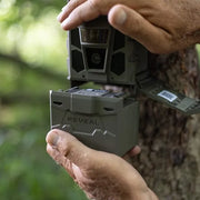 A close-up of a person's hands holding a gray REVEAL trail camera outdoors against a blurred green forest background and tree trunk.