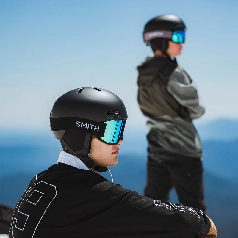 Two people wearing ski helmets with goggles against a mountainous background