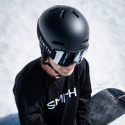 Person wearing a black Smith helmet and goggles with a snowboard in the background