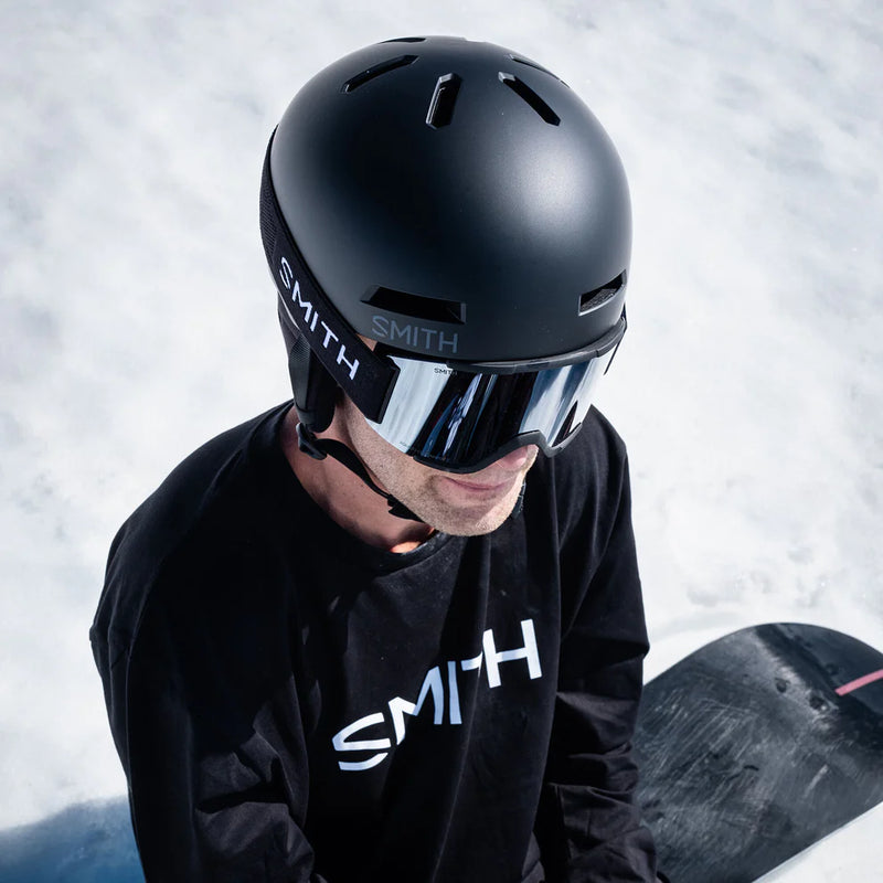 Person wearing a black Smith helmet and goggles with a snowboard in the background