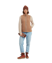 Person wearing a brown vest, beige sweater, light blue jeans, and red boots on a white background