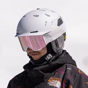 Person wearing a white helmet and goggles with a black jacket and colorful design, standing against a snowy background.