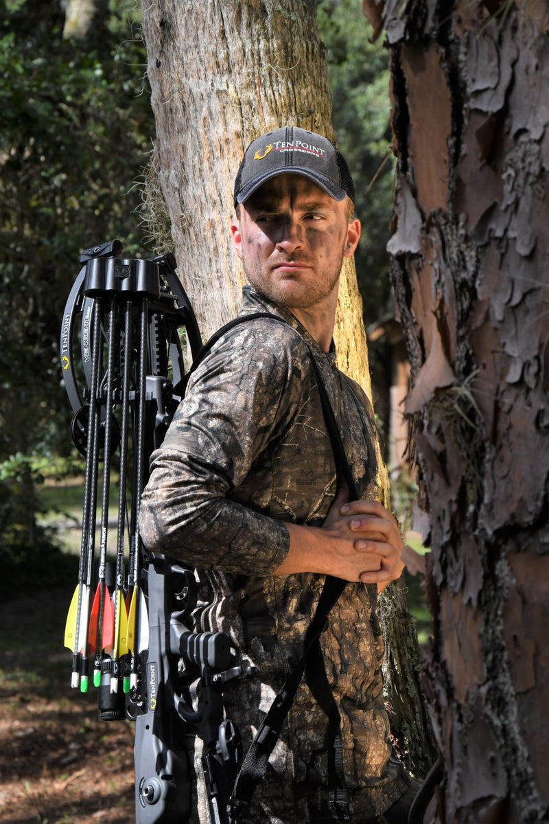 A man in camouflage hunting clothing and a cap stands behind a tree in a forest, holding a crossbow with arrows attached. 