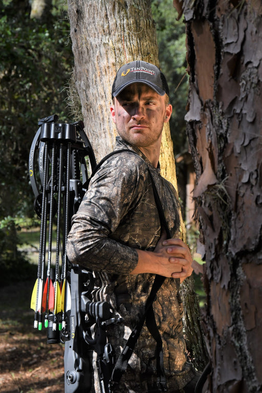 A man in camouflage hunting clothing and a cap stands behind a tree in a forest, holding a crossbow with arrows attached. 