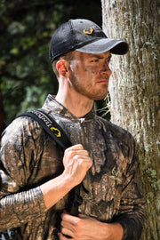 A man in camouflage hunting apparel stands beside a tree trunk outdoors. He is wearing a dark mesh-back cap with a yellow logo, a camouflage long-sleeve shirt, and has a black shoulder strap over his shoulder.