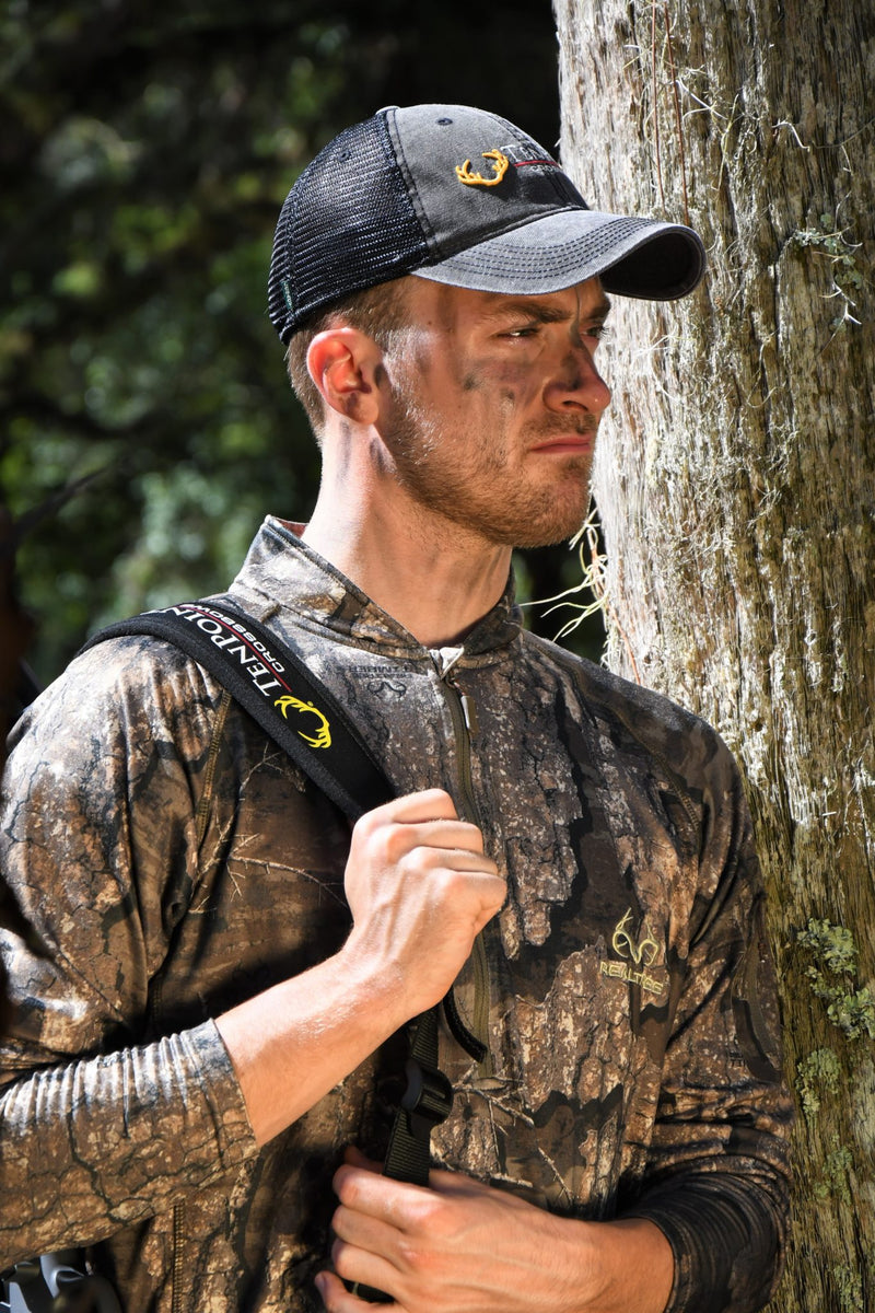 A man in camouflage hunting apparel stands beside a tree trunk outdoors. He is wearing a dark mesh-back cap with a yellow logo, a camouflage long-sleeve shirt, and has a black shoulder strap over his shoulder.