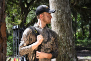 A man in a forest wearing a camouflage hunting shirt and a black cap with a yellow logo, carrying a bow on his back.