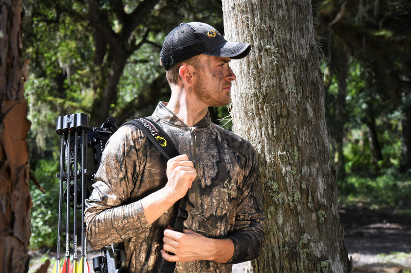 A man in a forest wearing a camouflage hunting shirt and a black cap with a yellow logo, carrying a bow on his back.